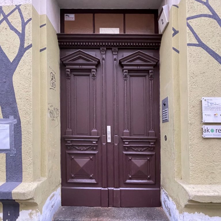 Restaurierte Eingangstür Braune Doppeltür mit dekorativen Elementen, umgeben von gelbem Putz und Baum-Motiven.